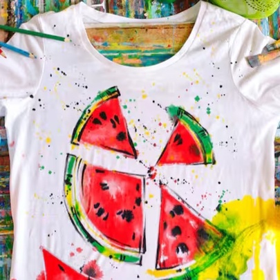 A white T-shirt painted with a watermelon slice design using fabric paint, with pots of paint and brushes on a craft table.