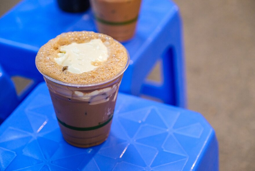 An iced coffee with foam and cream on top on a blue plastic stool.