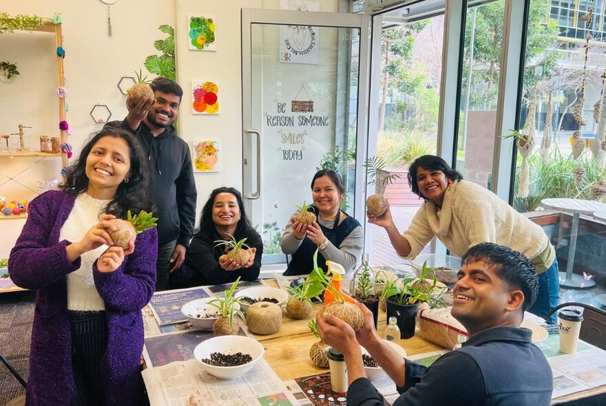 Smiling people at a workshop, the table covered in plants and soil, the participants making kokedama balls, in a bright and airy space.