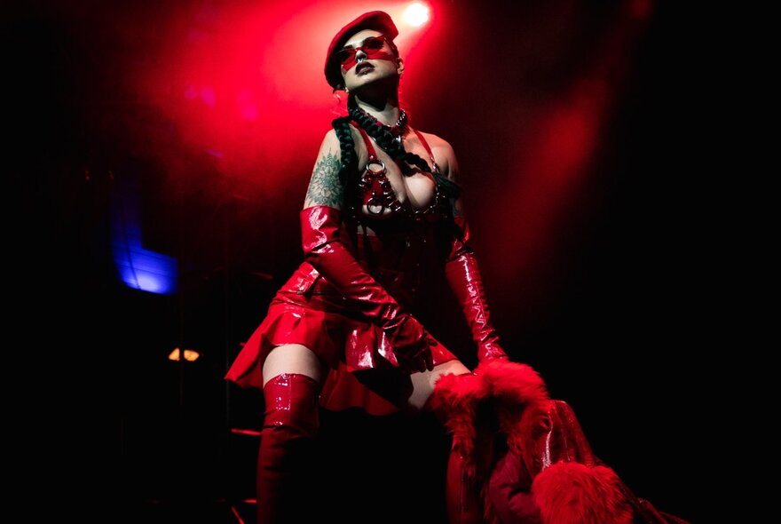 A burlesque dancer on stage wearing a red vinyl outfit and red glasses backlit by a red stage light. 