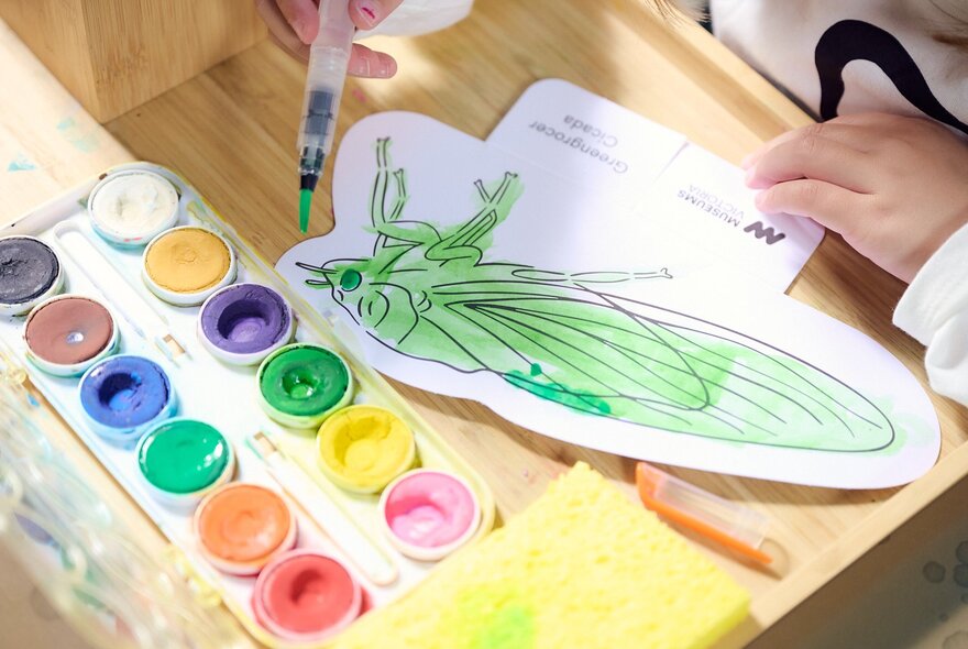 A child's hand painting a grasshopper cut-out using green watercolours, at an activity table at Melbourne Museum.