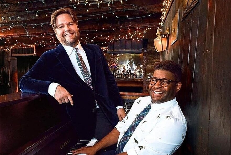 Two men smiling broadly, one seated in front of a piano, the other leaning on it. 