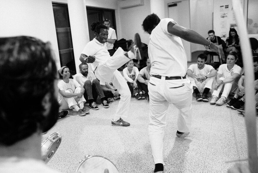 People participating in a Capoeira Angola class, with two people performing bold athletic moves in the centre of a seated group of onlookers. 