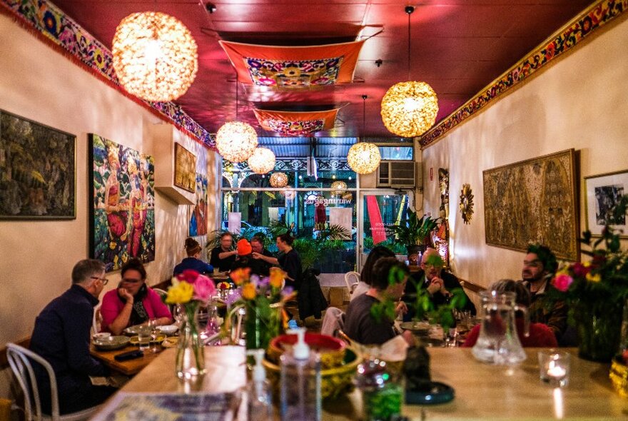 A group of people are sitting in a restaurant, there are lanterns hanging from the ceiling and Indonesian decorations on the walls and ceiling