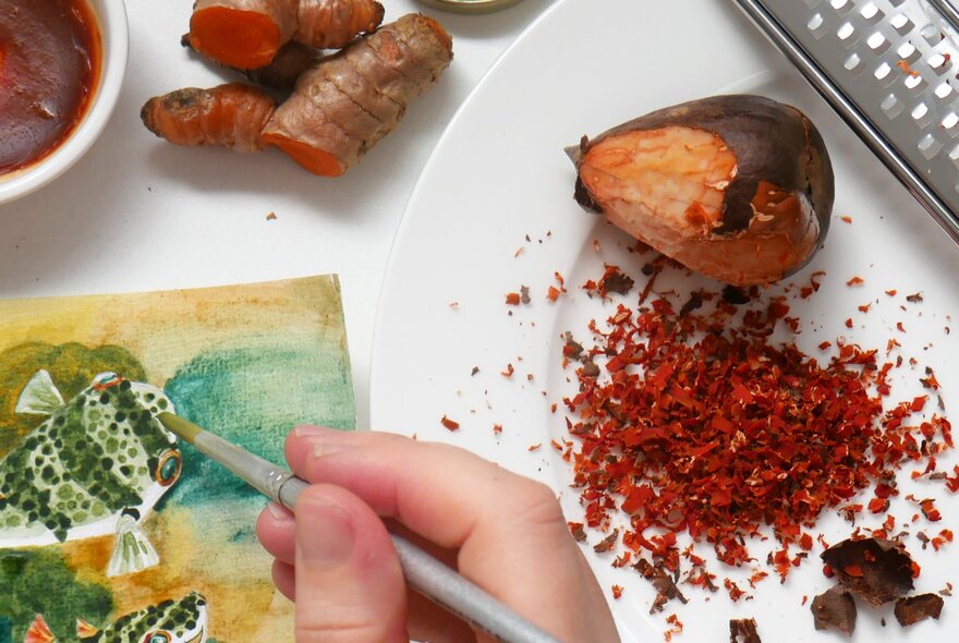 A hand using a fine brush to paint, with a plate of avocado pip gratings and ground fresh turmeric root used to make the coloured paints.
