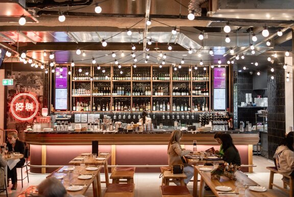 The festoon-lit interior of a rooftop bar with long tables and bench seats.