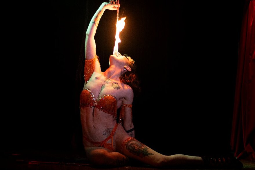 A tattooed woman in red lingerie holding a flaming fire-eating apparatus over her mouth.