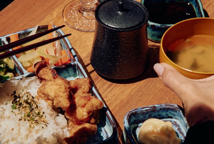 A blue earthenware bento box with rice, kariaage, pickles and chopsticks with a smaller blue earthenware dish with ginger and a miso soup in a bowl.