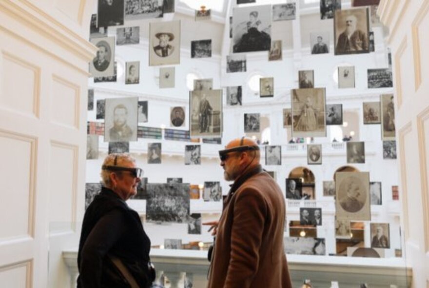 Two older people wearing mixed-reality headsets in front of a gallery wall with a crowded display of black and white portraits.