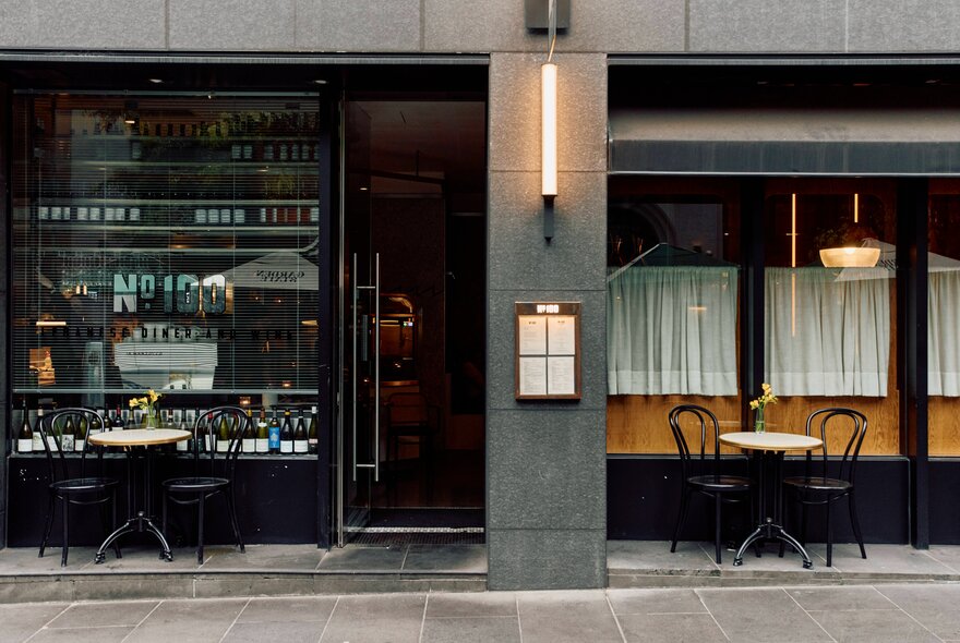 The exterior of No. 100 Flinders Lane wioth small tables out the front and a thin light on the pillar between buildings.
