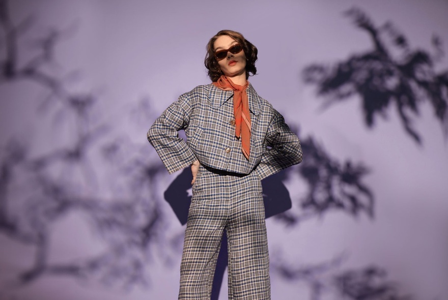 A model wearing a checked jacket and matching trousers, a tan scarf tied around her neck, a large leaf shadow on the wall in the background.