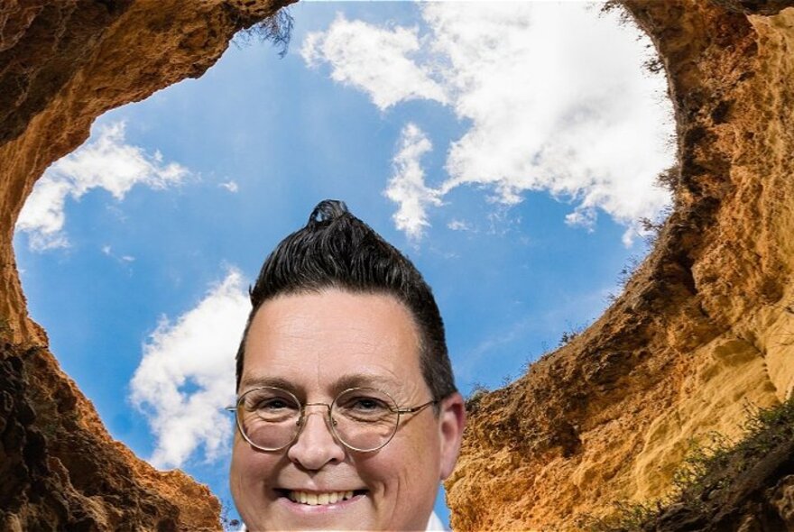 The smiling head of a person, wearing glasses, with a background of a hole dug into the clay earth and blue sky and clouds.