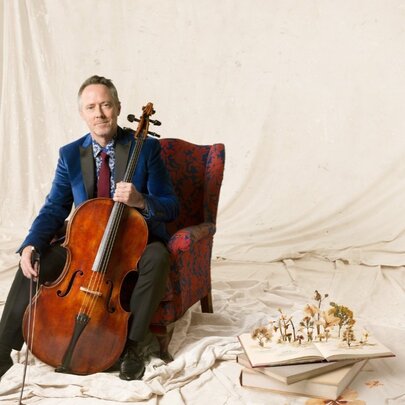 Musician seated against a backdrop of flowing white material, holding a cello.