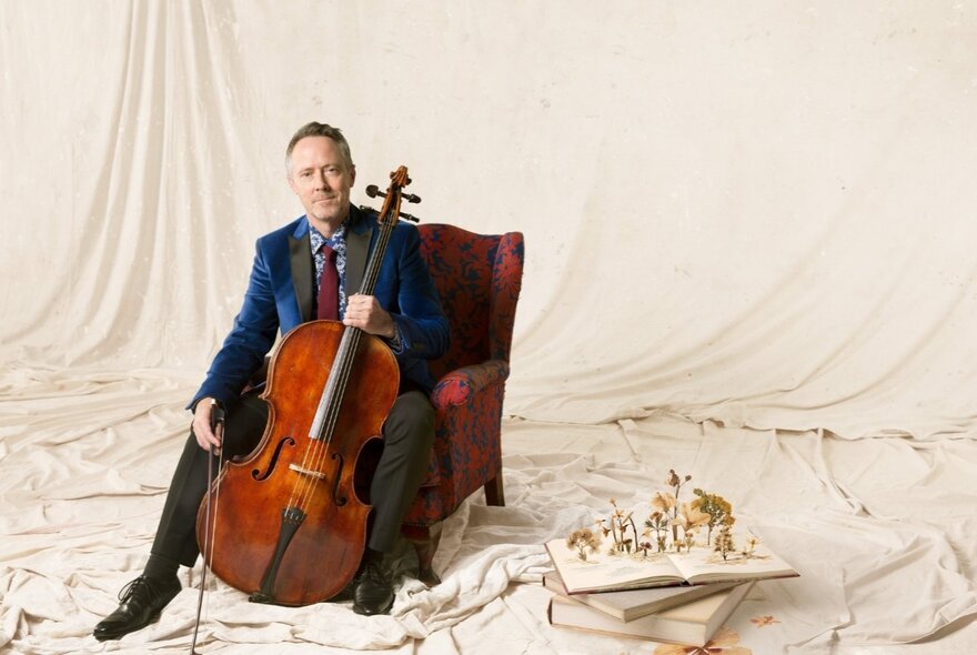 Musician seated against a backdrop of flowing white material, holding a cello.