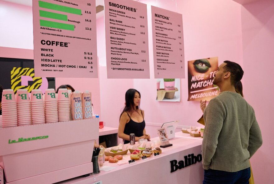 People looking up at the Balibola menu on the wall of Balibola cafe, which is painted nearly all in pale pink; ordering with the person serving behind the counter.