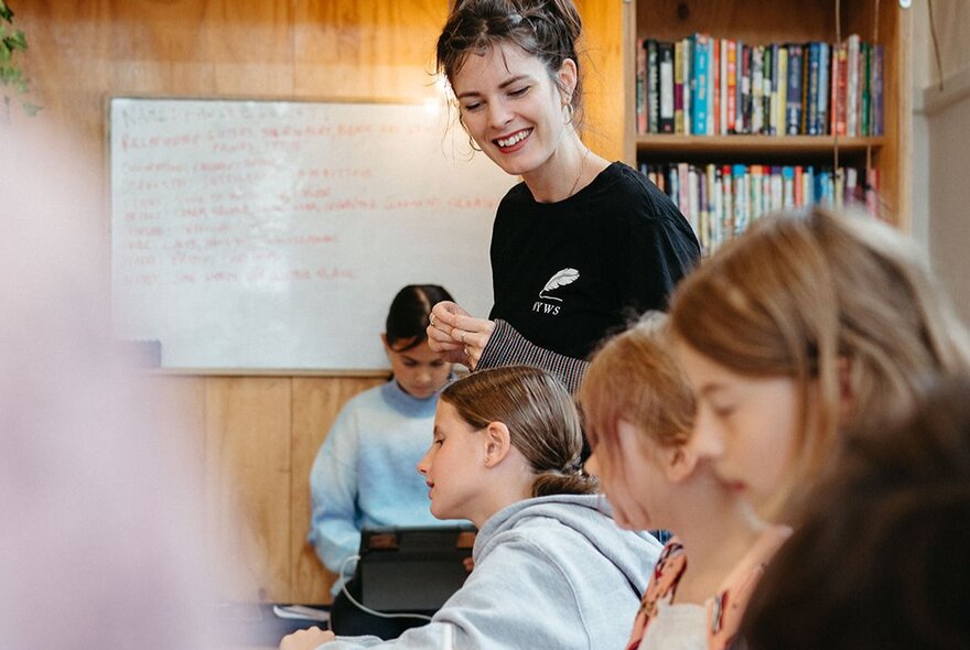 A teacher stands behind seated students writing at a table in a studio workshop, with bookshelves and a whiteboard in the background.