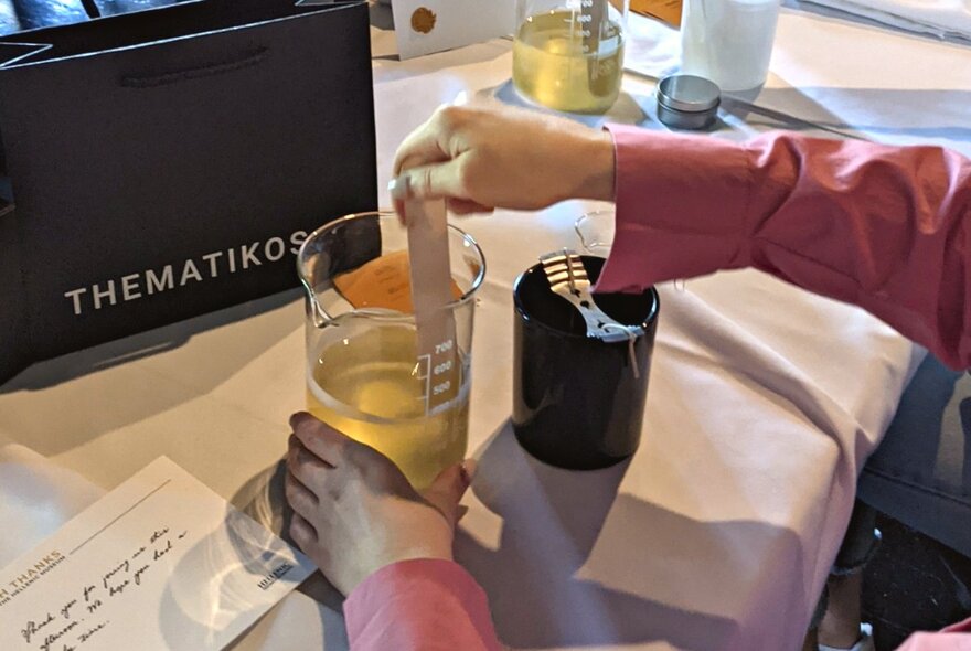 A table prepared with tools and ingredients ready for a candle making workshop at the  Hellenic Museum, with a hand mixing liquid in a glass beaker.