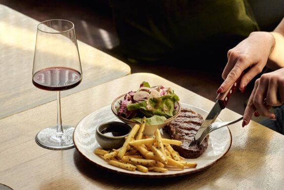 Person using a knife and fork to cut into a steak on a plate with French fries, salad, and a side sauce, on a table next to a glass of red wine.