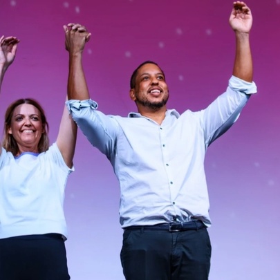 A woman and a man, both standing with hands in the air and smiling against a purple-hued background. 