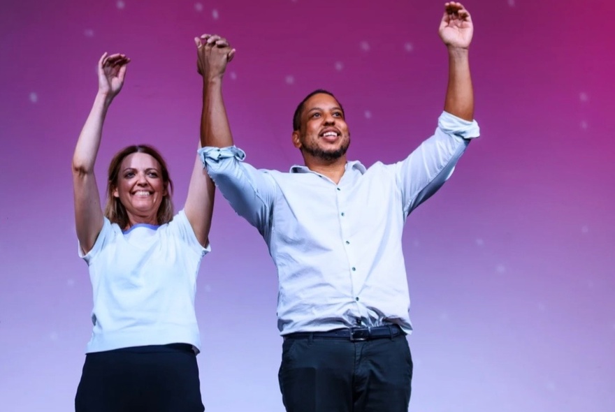A woman and a man, both standing with hands in the air and smiling against a purple-hued background. 