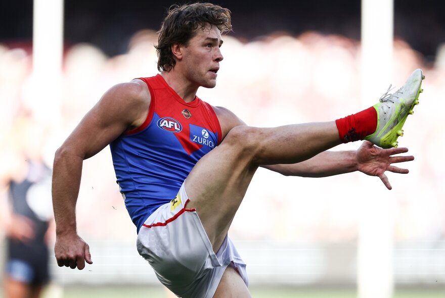 A Melbourne AFL football player with arms outstretched during a game and his leg raised high in the air after having kicked the football.