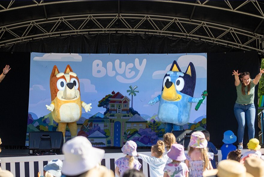 Children wearing sunhats watching a Bluey interactive show with stage and screen.