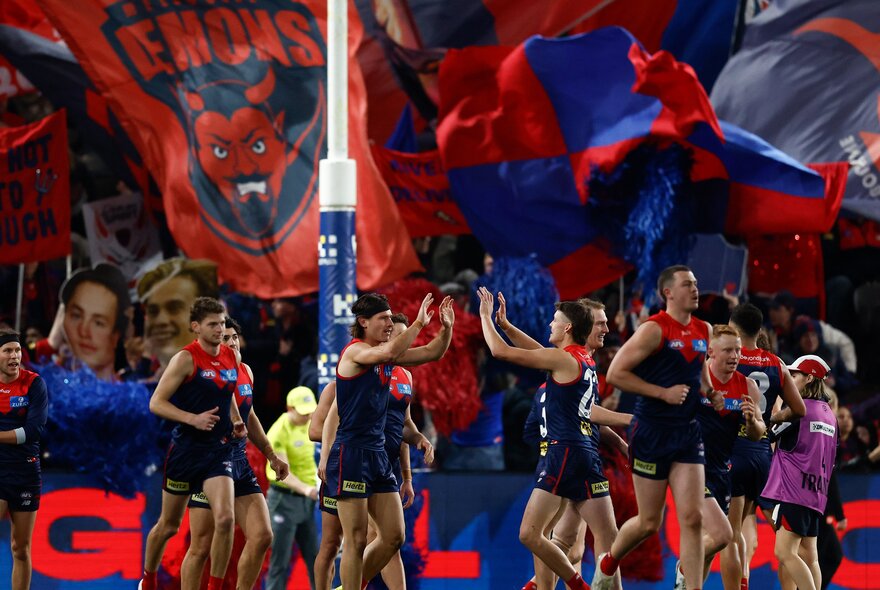 Melbourne AFL football players celebrating in front of the goal posts, their fans in the stands in the background waving banners and flags in their team's colours.