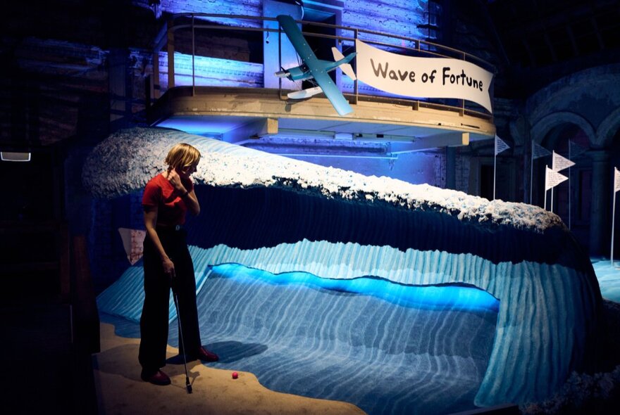 A mini golf putting hole shaped like a blue wave, a woman contemplating the hole and her ball.