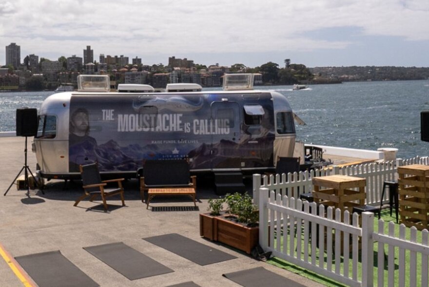 The Movember Airstream trailor on a concrete platform in front of the blue ocean with city skyline in the background.