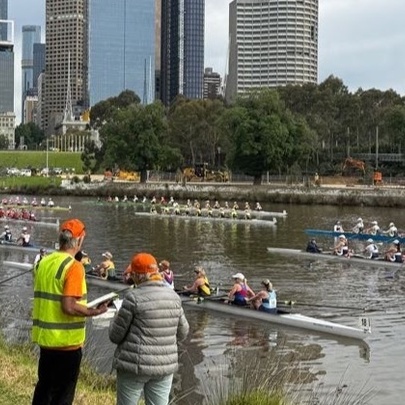 Team officials standing on the banks of the Yarra with competing rowers lined up on the water, city buildings in the background.