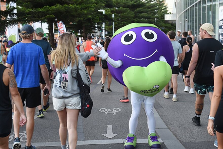 People walking around the race village of the Run for the Kids at District Docklands, with a mascot in costume high-fiving people as they walk past.