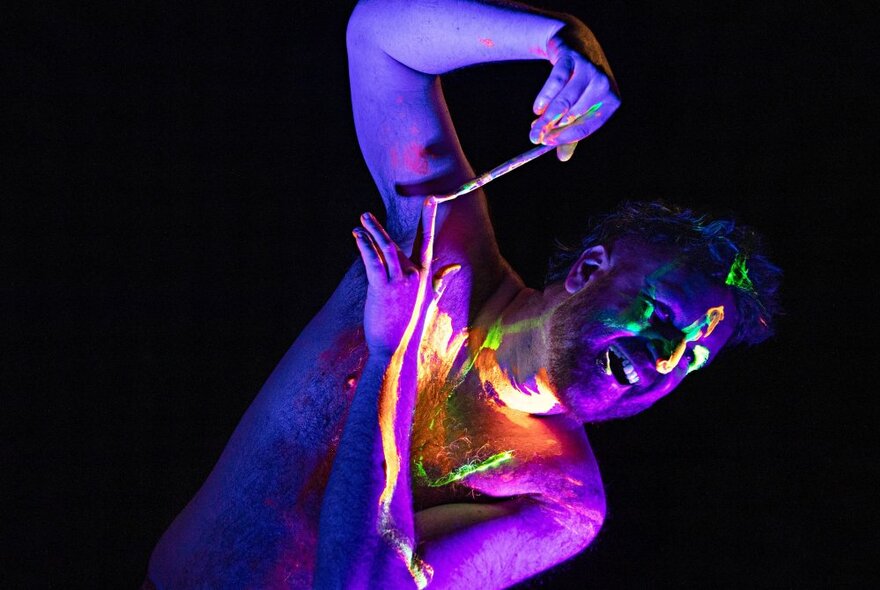 A man in the dark, painting himself with neon coloured paint that is glowing.