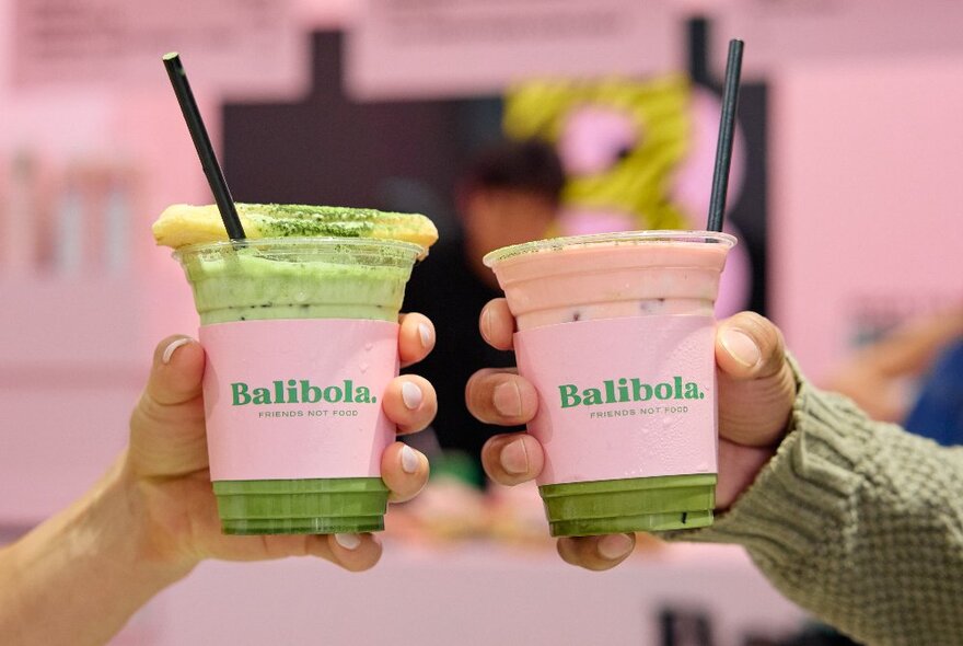 Two hands holding two green matcha drinks in plastic takeaway cups, the words Balibola written on the cups. 