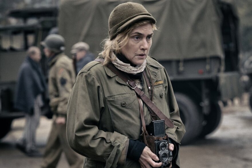 Actress Kate Winslet in the character of war photographer Lee Miller, holding a camera and wearing a khaki army coat and a hard helmet.