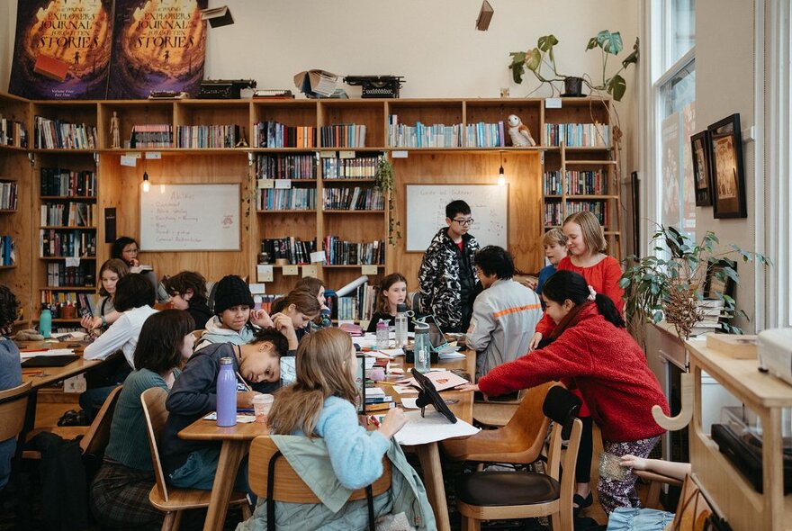 Young writers seated at tables writing in a workshop with papers and books.
