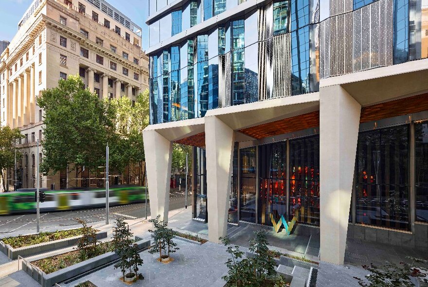 W hotel ground floor with postmodern concrete pillars supporting glassed upper floors, with flower beds and passing tram.