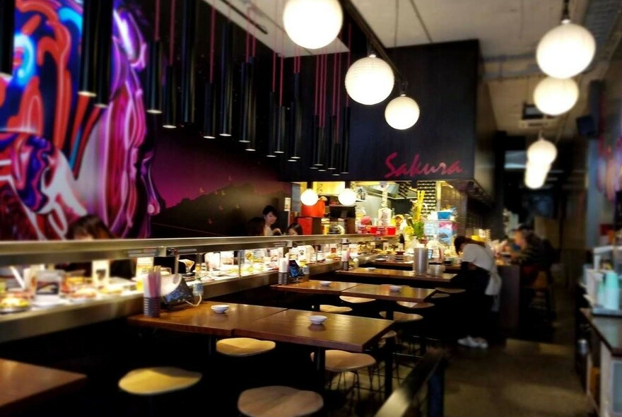 Restaurant interior with round pendant lights, dark tables, wall mural and backlit counter.