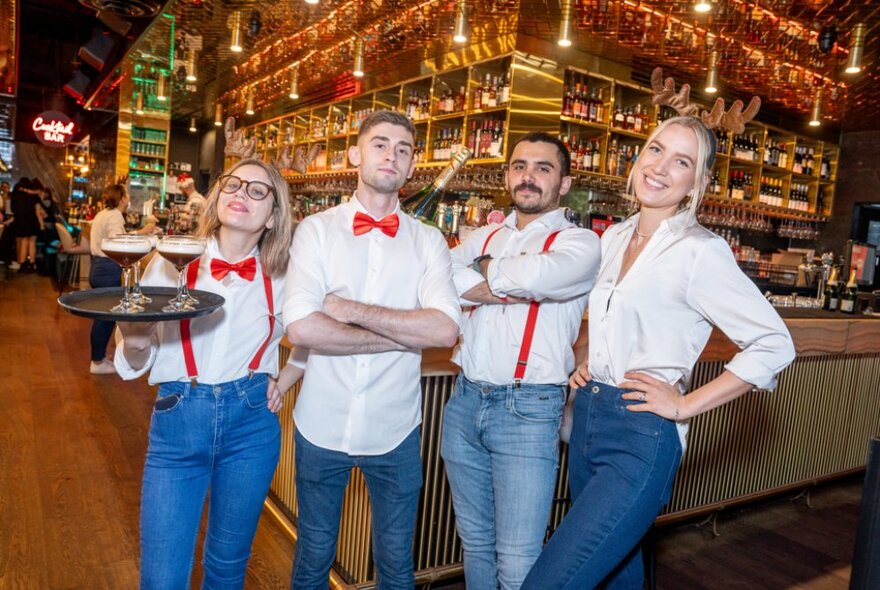 Four wait staff posting in front of a bar, some with red braces and bow ties. 