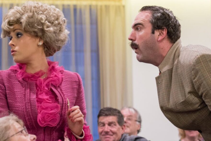 People dressed as characters from Fawlty Towers, Basil sticking his head forward as Sybil turns her head away, seated people in the background. 
