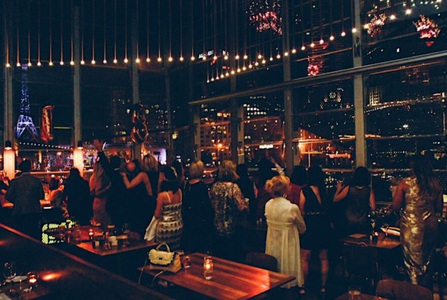 Dining patrons standing inside Taxi Kitchen restaurant, looking through the large windows surrounding them at views of the city at night.