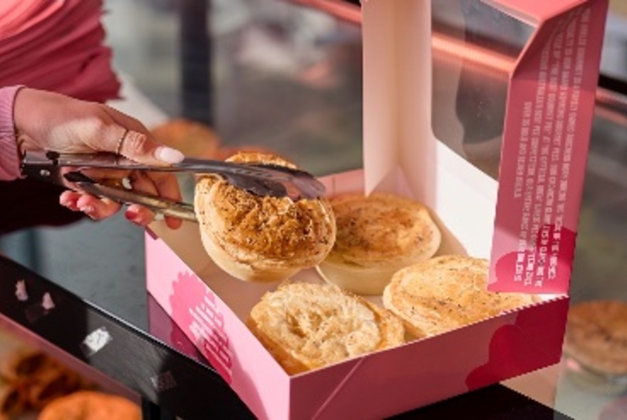 Three gourmet pies in a pink cardboard display box, with a woman holding a fourth in a pair of tongs.