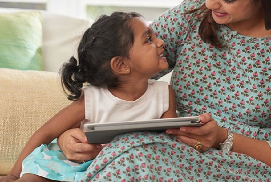 A mother and child seated on a couch close together looking at each other, a story book on the mother's lap.