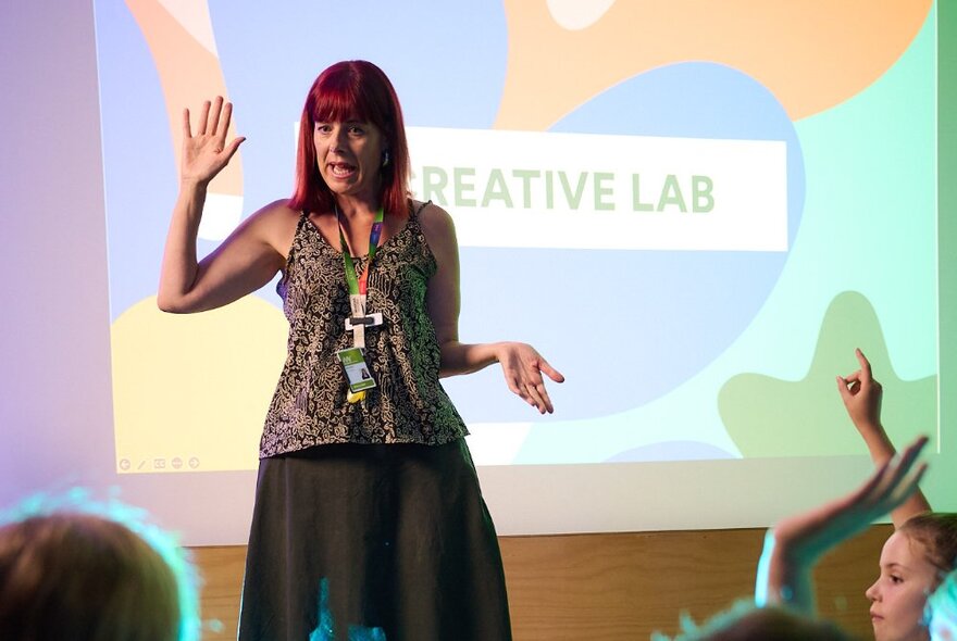 A teacher in front of a classroom with her hand up and speaking in front of a large projection screen. 