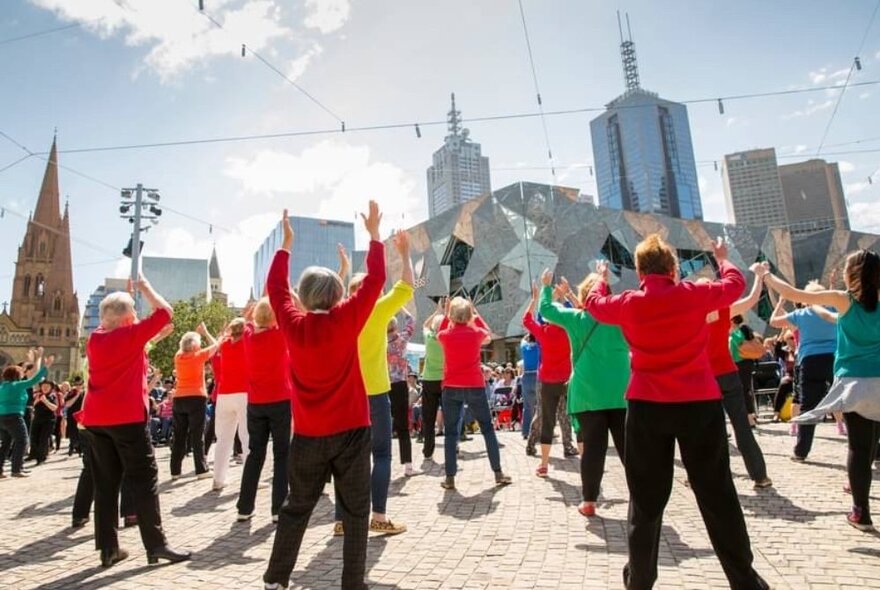 Looking at the back of a group of people in red, yellow, blue and green all with their hands in the air in an outdoor class at Federation Square. 