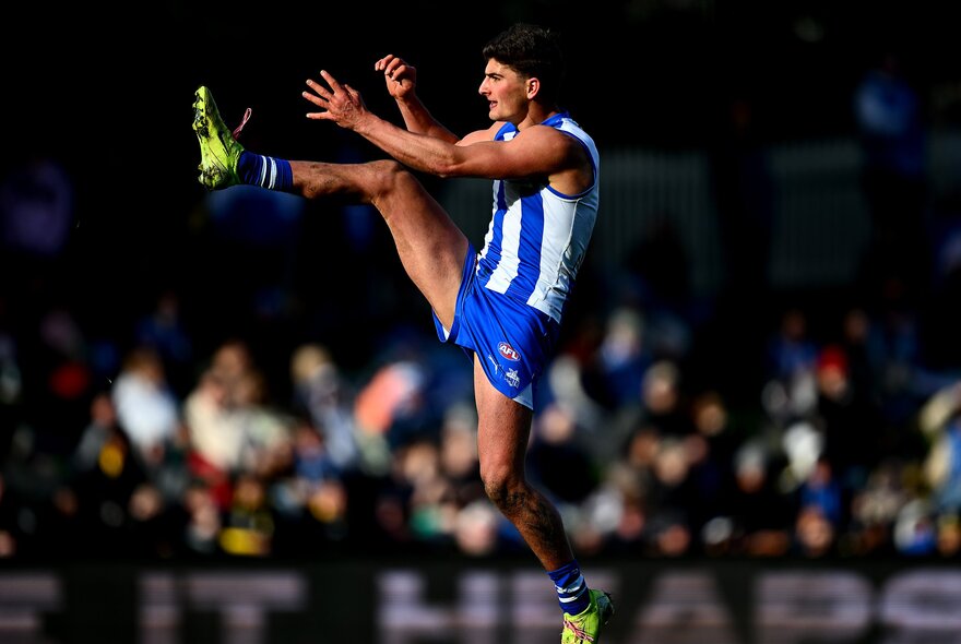 A North Melbourne AFL player in mid air with his leg stretched high after having kicked a football.