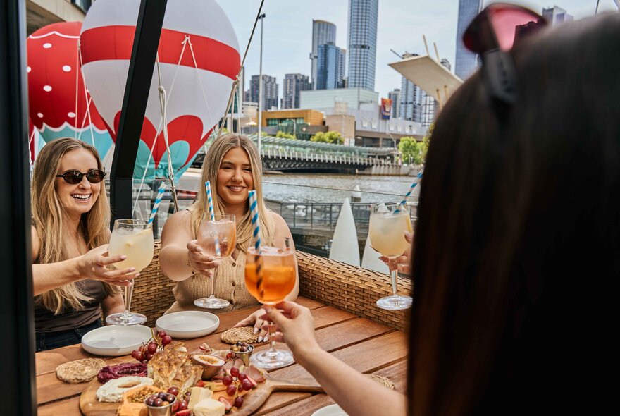 Three friends eating and drinking at a table an outdoor dining booth with a mini hot air balloon suspended above them and balloons visible in the background, with The Yarra River and city buildings in the background.