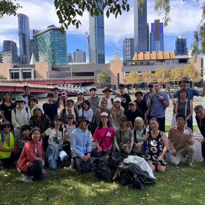 A large group of community volunteers gathered on a grassy bank along the Yarra River in Melbourne, posing with bags of collected rubbish after a riverside clean-up event.