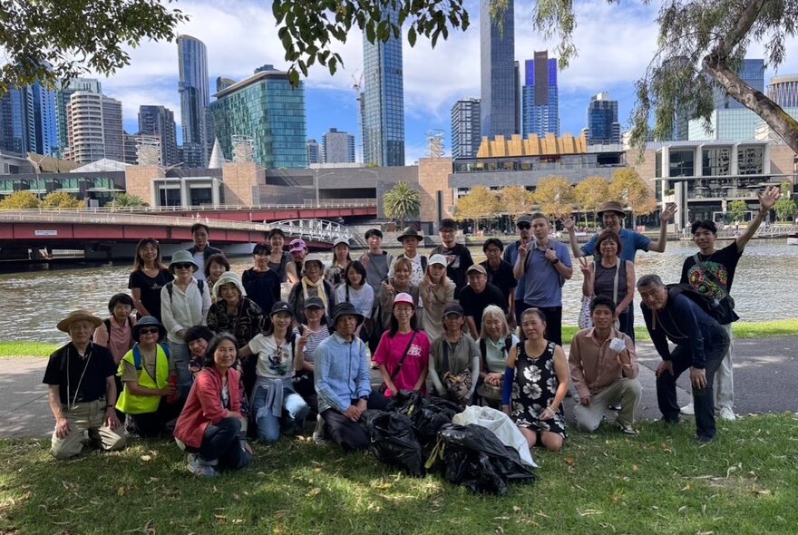 A large group of community volunteers gathered on a grassy bank along the Yarra River in Melbourne, posing with bags of collected rubbish after a riverside clean-up event.