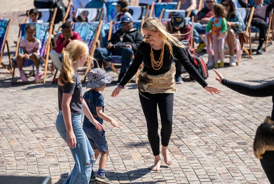 A dancer interacting with two young children and dancing with them at Fed Square's main outdoor plaza, with people seated on deck chairs in the background.