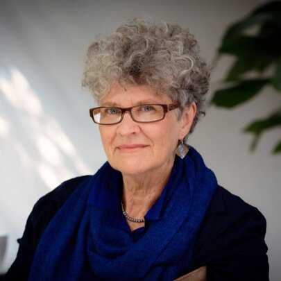 Author Kate Grenville, with grey hair and spectacles, in a shady room with plants.